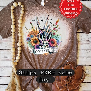 May include: Brown t-shirt with a bleached design featuring a skeleton hand making a peace sign, surrounded by colorful flowers and the words "GOOD VIBES." The shirt also has the text "Ships FREE same day."