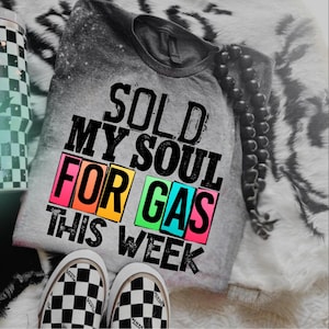May include: A gray t-shirt with the text "SOLD MY SOUL FOR GAS THIS WEEK" in black, with colorful block letters. The shirt is paired with black and white checkered Vans sneakers. A black beaded necklace and a checkered tumbler are also visible.