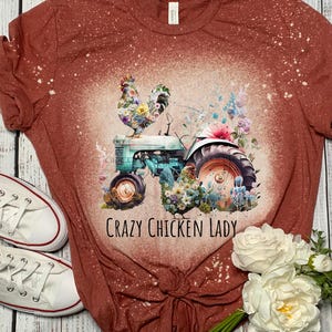 May include: A rust-colored t-shirt with a floral rooster design on a vintage tractor. The text "Crazy Chicken Lady" is printed below the image. White sneakers and flowers are also in the image.