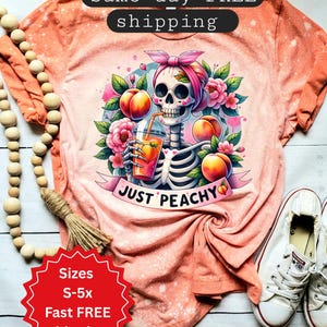 May include: Peach-colored t-shirt with a graphic of a skeleton wearing a pink bow and holding a peach drink. The design includes peaches and flowers, with the text "JUST PEACHY" on a banner. The shirt has a bleached effect.
