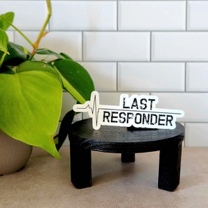 May include: White sticker with black text and a black heartbeat graphic that reads "LAST RESPONDER". The sticker is placed on a black round pedestal.