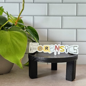 May include: A black table with a sticker that says "FORENSICS" in white letters. The sticker is made up of different images related to forensics, such as a magnifying glass, a ruler, and a fingerprint.