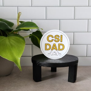 May include: White round sticker with the text "CSI DAD" in yellow letters. The sticker is on a black wooden pedestal.