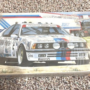 Puede incluir: Una caja con una ilustración de un coche de carreras BMW blanco, con detalles en azul, rojo y negro y logotipos de patrocinadores. El coche tiene el número 5 en el lateral. La caja también muestra un camión y las palabras "Original BMW Teile".