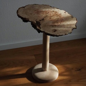 Op de afbeelding: Een houten bijzettafel met een blad van natuurlijk hout en een lichte houten basis. Het blad is een dikke plak hout met een natuurlijke rand en een uniek nerfpatroon. De basis heeft een eenvoudige cilindrische vorm met een afgeronde basis.