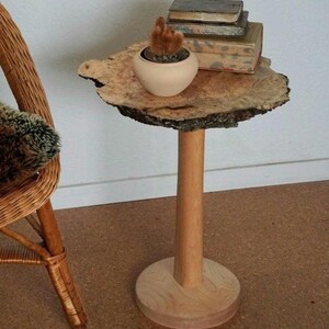 Op de afbeelding: Een houten bijzettafel met een naturel houten blad en een enkel, rond, houten poot. Het blad is gevormd als een boomschijf en heeft een natuurlijke schorsrand. Een kleine plant in een pot en een stapel boeken staan op de tafel.