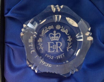 May include: A clear, faceted glass ashtray with a blue interior. The ashtray features the initials "ER" with a crown, floral designs, and the dates "1952-1977" and "1977". It is presented in a black box with a blue satin lining.