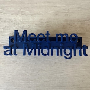 Puede incluir: Un letrero de plástico azul con el texto "Meet me at Midnight" en letras blancas y negritas.
