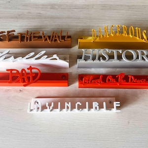 Könnte beinhalten: Ein Satz von 8 Wandplatten mit Albumtiteln des Künstlers Michael Jackson. Die Platten sind aus weißem, grauem, braunem und rotem Kunststoff. Die Titel sind "Off the Wall", "Thriller", "Bad", "Dangerous", "History", "Blood on the Dance Floor" und "Invincible".