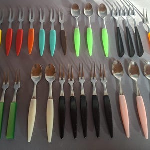 Puede incluir: Colección de cubiertos vintage, que incluye cucharas y tenedores, con mangos coloridos en amarillo, rojo, naranja, turquesa, marrón, verde, negro y rosa. Los utensilios de acero inoxidable están dispuestos sobre una superficie gris.