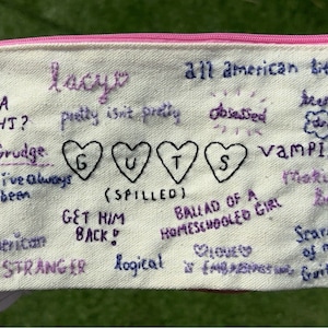 May include: A white canvas zippered pouch with a pink zipper. The pouch is embroidered with song titles and lyrics from the album "GUTS" by Olivia Rodrigo. The song titles include "Bad Idea Right?", "Pretty Isn't Pretty", "All American Bitch", "Obsessed", "Vampire", "Making the Bed", "Get Him Back", "Ballad of a Homeschool Girl", "Stranger", "Logical", "Scared of My Guitar", and "The Grudge".