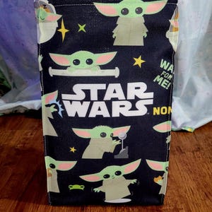 Pu&ograve; includere: Una borsa rettangolare nera a tema Star Wars, con illustrazioni di Grogu e il logo di Star Wars. La borsa presenta un motivo ripetuto di Grogu, stelle e il testo "WA FOR ME!"