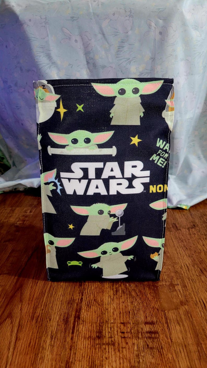 Pu&ograve; includere: Una borsa rettangolare nera a tema Star Wars, con illustrazioni di Grogu e il logo di Star Wars. La borsa presenta un motivo ripetuto di Grogu, stelle e il testo "WA FOR ME!"