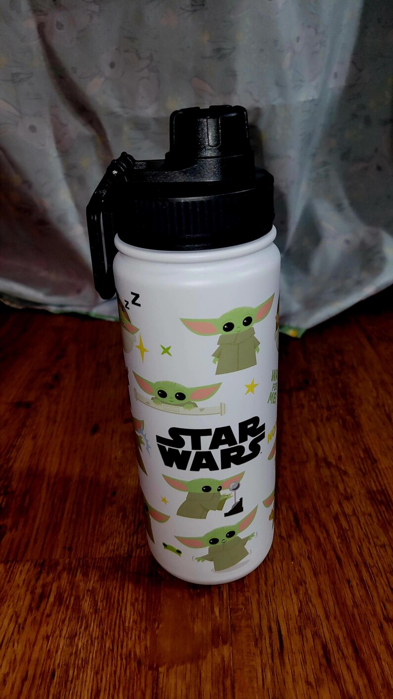 Pu&ograve; includere: Borraccia bianca con coperchio e manico neri. La borraccia &egrave; decorata con immagini di Grogu di Star Wars, insieme al logo di Star Wars. Il design include elementi verdi, gialli e bianchi.