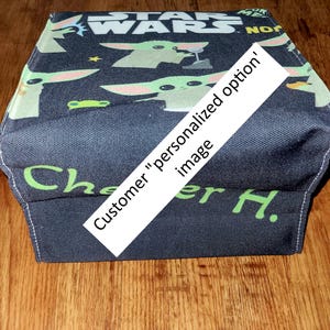 Pu&ograve; includere: Una mascherina blu scuro a tema Star Wars con Grogu e il logo di Star Wars in bianco. La mascherina ha un testo verde che dice "Che" e "er H.". Un banner diagonale bianco dice "Customer 'personalized option' image".