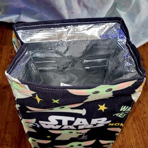 Pu&ograve; includere: Borsa termica per il pranzo con sfondo nero con la scritta "STAR WARS" e immagini di Grogu. La borsa ha un interno argentato isolato e un bordo nero. La borsa &egrave; aperta, rivelando l'interno.