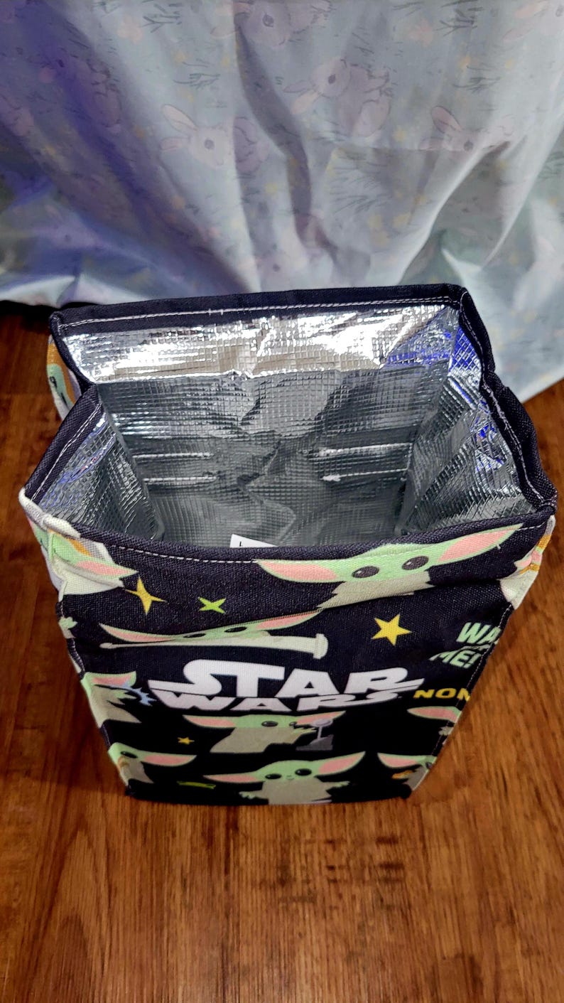 Pu&ograve; includere: Borsa termica per il pranzo con sfondo nero con la scritta "STAR WARS" e immagini di Grogu. La borsa ha un interno argentato isolato e un bordo nero. La borsa &egrave; aperta, rivelando l'interno.