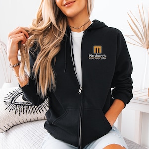 May include: Black zip-up hoodie with the "Pittsburgh Trauma Medical Center" logo in gold and white on the upper left chest. The hoodie has a full zipper, a hood, and front pockets. The person is wearing a white t-shirt underneath.