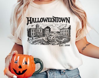 May include: Cream-colored t-shirt featuring a black and white graphic of a town scene with the text "Halloweentown" and "EST. 1998". An orange pumpkin-shaped mug with a carved face is held in the foreground. The mug has a green handle.