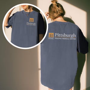 May include: A navy blue t-shirt with the text "Pittsburgh Trauma Medical Center" printed on the front and back. The logo is a stylized orange and yellow archway. The shirt is a comfortable, casual style.