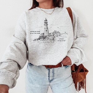 May include: A light gray sweatshirt featuring a black and white illustration of a lighthouse on a rocky coast. The text on the sweatshirt reads "wanting was enough for me it was enough" and "to live for the hope of it all."