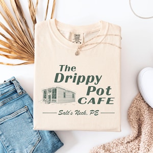 May include: A beige t-shirt with a green graphic design that says "The Drippy Pot Cafe - Salt's Neck, PE". The graphic features a small house with a snowy roof.