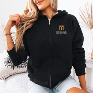 May include: Black zip-up hoodie with the text "Pittsburgh Trauma Medical Center" in gold lettering. The hoodie has a full zipper and a hood. The logo is a gold square with a stylized building.