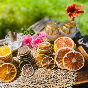 May include: A wooden tray containing a selection of dried citrus fruit slices, including orange, grapefruit, and lime. The fruit slices are stacked and surrounded by a beige, woven mat. Two gold-coloured containers with lids are in the background.