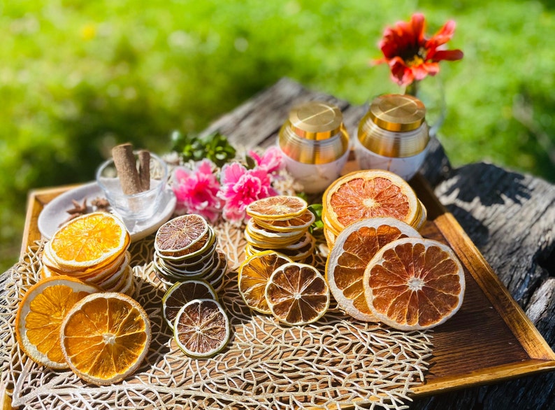 May include: A wooden tray containing a selection of dried citrus fruit slices, including orange, grapefruit, and lime. The fruit slices are stacked and surrounded by a beige, woven mat. Two gold-coloured containers with lids are in the background.