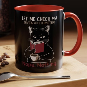 Könnte beinhalten: Schwarze Kaffeetasse mit rotem Rand. Die Tasse zeigt eine schwarze Katze, die ein Buch liest, mit dem Text "Let me check my giveashitometer Nope, Nothing".