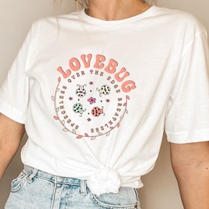Pode incluir: Camiseta branca com um gráfico circular rosa com o texto "LOVEBUG OVER THE EDGE BREATHLESS" e um design de quatro joaninhas com detalhes em rosa, verde e preto.