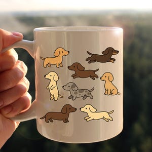 May include: A white ceramic mug decorated with cartoon dachshund illustrations in various poses and colours, including tan, brown, and grey. The mug has a curved handle and is held against a blurred green background.
