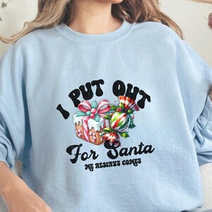 May include: Light blue sweatshirt with a Christmas-themed graphic that says "I Put Out For Santa He Always Comes". The graphic features a wrapped gift box with a red bow and candy canes.