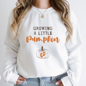 May include: White sweatshirt with the text "Growing a Little Pumpkin" in brown lettering. A small orange pumpkin with brown stem and two orange footprints is below the text.