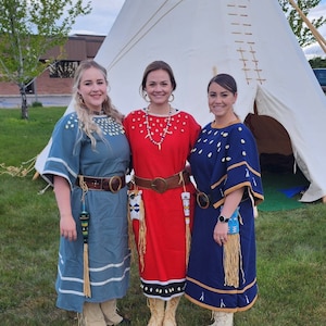 May include: Three women wearing traditional Indigenous clothing stand in front of a white teepee. The women are wearing blue, red, and navy blue dresses with beaded accents and fringe. They are smiling and looking at the camera.