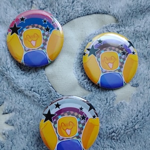 Puede incluir: Tres botones redondos con un diseño de personaje de dibujos animados. Los botones presentan un personaje amarillo con una cara sonriente, ropa azul y un arco iris con estrellas negras. El fondo es una tela azul claro y suave.