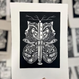 May include: A black and white linocut print depicting a central syringe design, with symmetrical anatomical illustrations on either side. The artwork is on white paper with a deckled edge. Other prints are visible in the background.