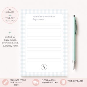 Può includere: Un blocco note a strappo A5 con motivo a quadretti vichy azzurro e bianco, composto da 50 pagine. Il blocco presenta linee orizzontali e il testo minor inconveniences department in alto. Una penna a sfera verde chiaro è posizionata a destra.