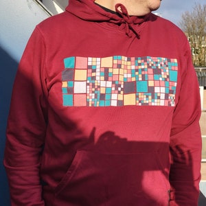 Puede incluir: Una sudadera con capucha roja con un estampado geométrico de colores en la parte delantera. El estampado está formado por cuadrados de varios tonos de rojo, amarillo, verde y azul.