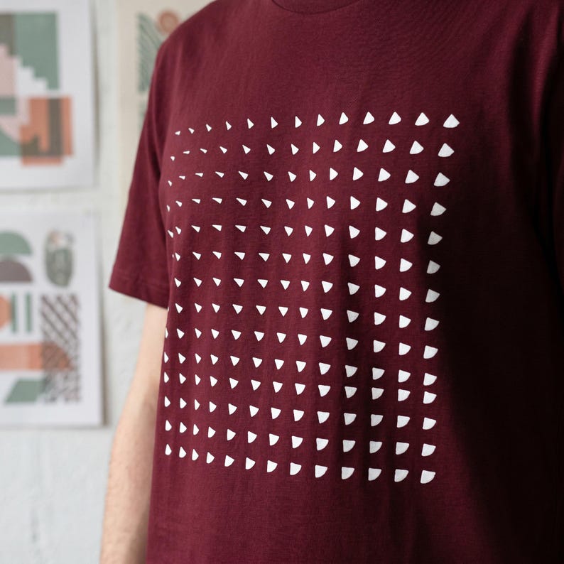 Close-up of a burgundy t-shirt with the generative white triangle grid print, worn in a studio with art prints on the wall