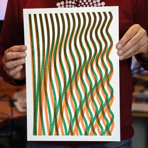 Person holding a handmade A3 risograph print with wavy green, orange and brown lines on white paper, showing the actual size of the abstract wall art