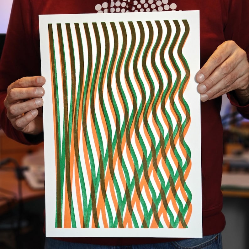 Person holding a handmade A3 risograph print with wavy green, orange and brown lines on white paper, showing the actual size of the abstract wall art