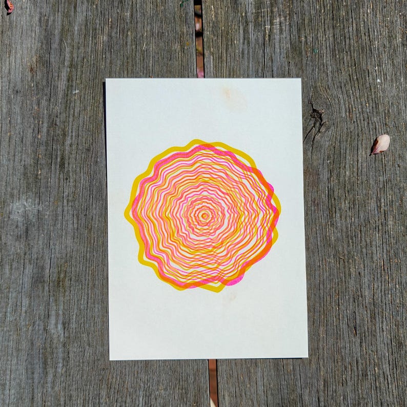 Top view of the A5 riso print on grey wooden planks, concentric tree rings in pink and yellow