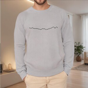 Puede incluir: Una sudadera gris claro con un diseño de línea negra en el pecho. La parte superior de manga larga con cuello redondo está hecha de un material suave. La sudadera se combina con pantalones beige.