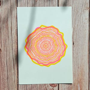 The A5 riso print lying flat on weathered wooden boards, concentric tree rings in pink and yellow