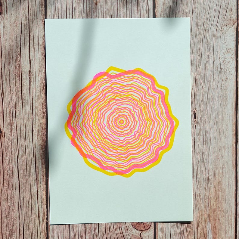 The A5 riso print lying flat on weathered wooden boards, concentric tree rings in pink and yellow