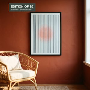 Framed A3 riso print with teal stripes and a fluorescent orange circle, in a black frame on a terracotta wall next to a rattan armchair