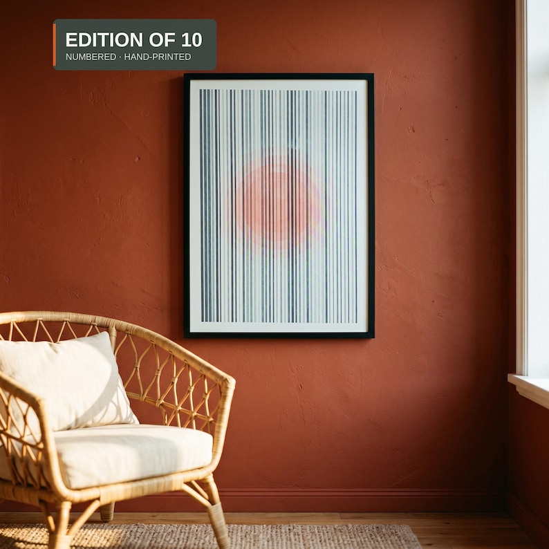 Framed A3 riso print with teal stripes and a fluorescent orange circle, in a black frame on a terracotta wall next to a rattan armchair