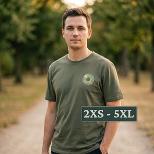 Può includere: Una t-shirt a maniche corte color verde oliva con un design grafico circolare sul petto sinistro caratterizzato da linee radiali gialle e blu. Un'etichetta rettangolare nell'angolo in basso a destra mostra il testo 2XS - 5XL in bianco.