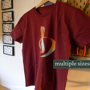 Könnte beinhalten: Ein maroonfarbenes T-Shirt mit einem farbenfrohen abstrakten Design. Das Design ist ein stilisierter Buchstabe "D" mit einem Farbverlauf von Orange, Gelb und Blau. Der Text "multiple sizes" ist am unteren Rand des Bildes sichtbar.